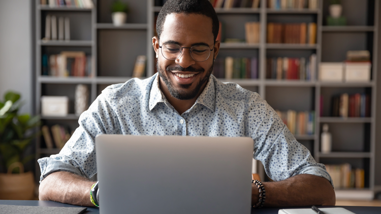 A man wearing a blue shirt smiles as he looks at his laptop.