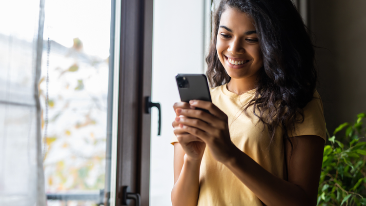 A woman holds a mobile phone and smiles as she looks at the screen.