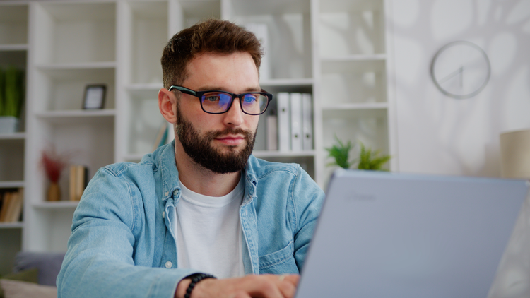 A man sits at a table and look at his laptop. He is wearing glasses and a denim shirt.