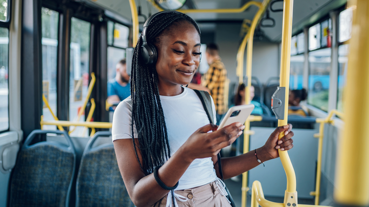 A young woman stands on a bus while looking at her phone. She is wearing headphones and smiling.