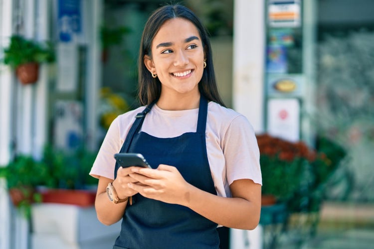 A person with long brown hair in a center part and gold hoop earrings and wearing pink t-shirt and black apron holds a smart phone and gazes smilingly to the top right corner of the image.