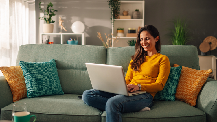 A woman sits on a sofa with a laptop on her lap. She is wearing a yellow jumper.