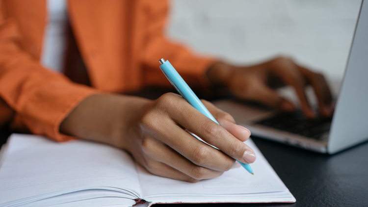 A hand holding a blue pen rests on an open notepad.