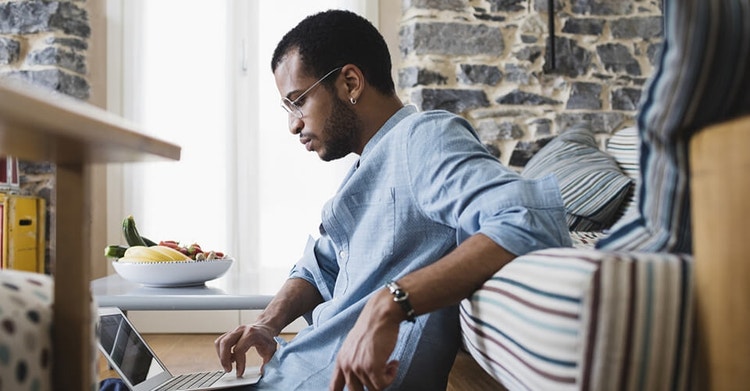 Man wearing glasses and a blue shirt sitting on the floor by his couch while on his laptop