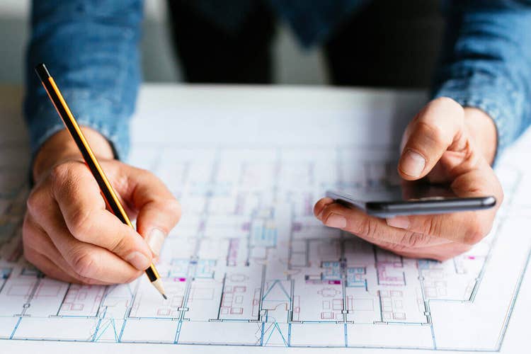 A close-up photo of a person's arms resting on a blueprint on a table. One hand is holding a pencil and the other hand a smartphone.