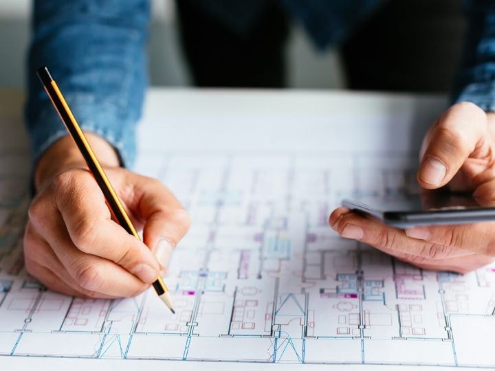 A close-up photo of a person's arms resting on a blueprint on a table. One hand is holding a pencil and the other hand a smartphone.