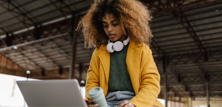 Personne, casque autour du cou et tenant un mug de voyage, les yeux rivés sur l’écran de son ordinateur portable