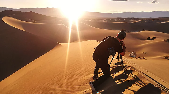 A photographer setting up a camera on a tripod on top of a desert dune while the sun sets in the background