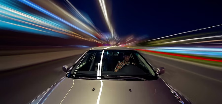 Un homme au volant d’une voiture gris métallisé fonce dans une rue tandis que des lignes floues aux couleurs vives défilent à toute vitesse en arrière-plan.