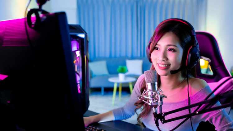 Woman live streaming A woman sits at a desk. She is wearing headphones and has a mic on the desk in front of her.