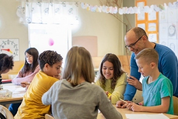 Right: A teacher looking over the work of four students who are sitting around a large desk.