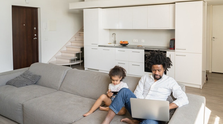 A parent sitting on a couch next to their child and signing a document on their Mac laptop