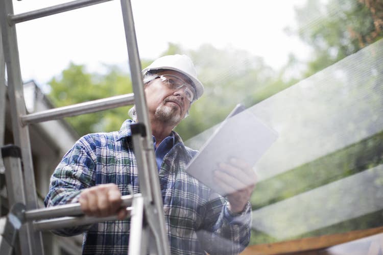 A roof contractor holding a tablet device while standing on a ladder