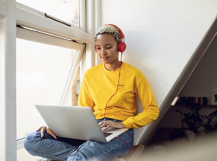 A college student listening to music and filling out a form on their laptop beside an open window