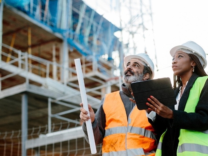 A contractor and client dressed in PPE looking over a construction bid template and blueprints while on the job site