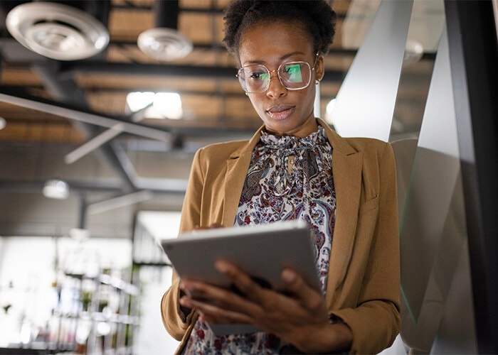 An office worker leaning against a wall while going over a digital form on their tablet