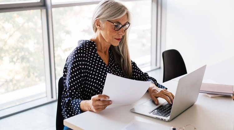 A landlord sitting at a desk reviewing a printed apartment lease agreement while using their laptop