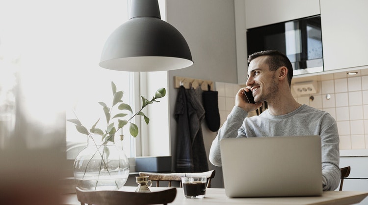 A person talking on the phone while sitting at a table with a laptop and a cup of coffee