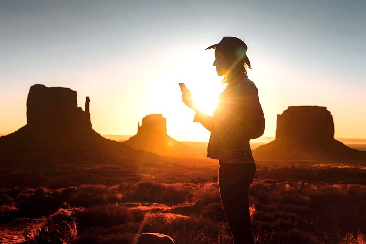 Sunset silhouette of person wearing cowboy hat looking at phone with Monument Valley rock formations in the distance