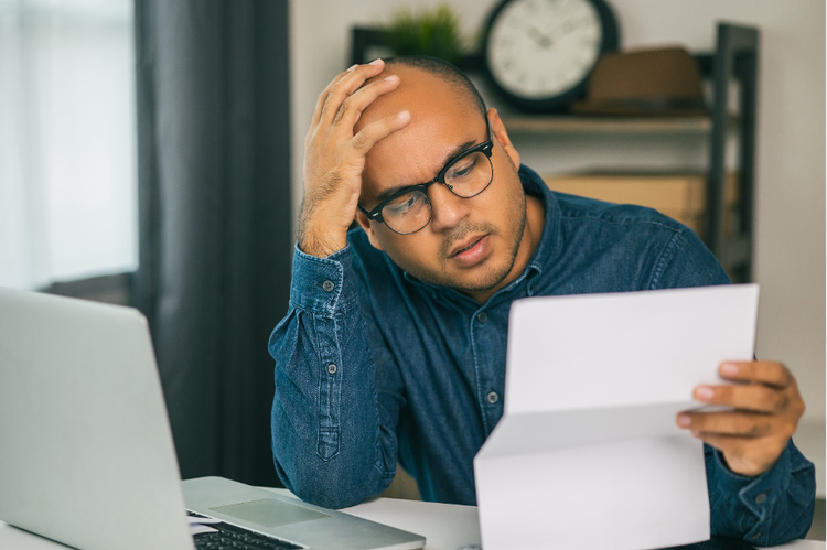 Man sitting in front of laptop, holding an invoice, looking puzzled at its contents.