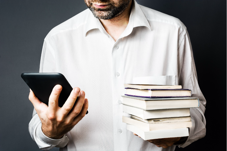 A man reads on a Kindle in one hand while holding a stack of books in the other.