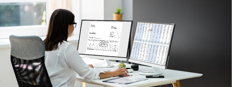 A woman sits at a desk with two computer monitors showing invoices and financial data.