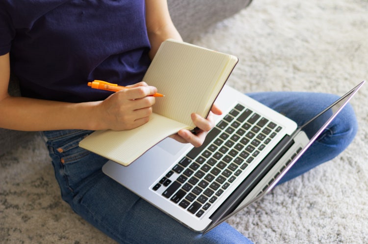 una ragazza seduta su un tappeto con un laptop appoggiato sulle gambe scrive appunti su un quaderno