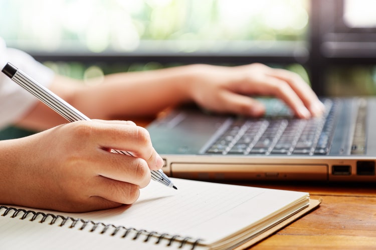 Una mano in primo piano che scrive su un quaderno mentre l'altra mano in secondo piano digita sulla tastiera di un computer portatile.