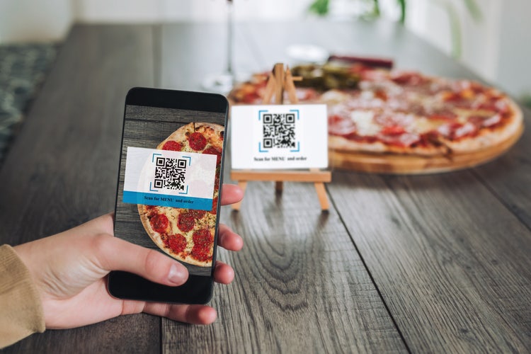 Smartphone che inquadra il QR code con il menu di un ristorante, con una pizza sullo sfondo.