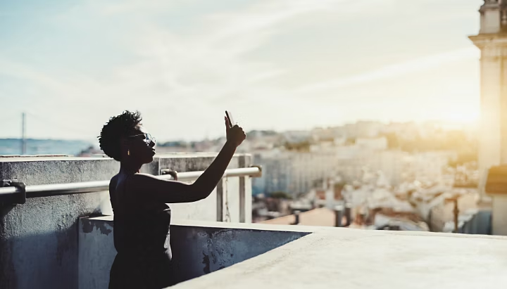 A-silhouette-of-a-young-black-woman-in-eyeglasses-taking-pics-on-her-cellphone-on-the-roof-with-a-sunny-cityscape-around-her-an-african-female-is-making-a-selfie-via-a-smartphone-on-the-rooftop