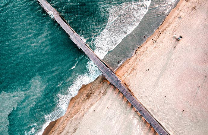 Drone image of sunny beach and ocean with a wooden pier in the center.