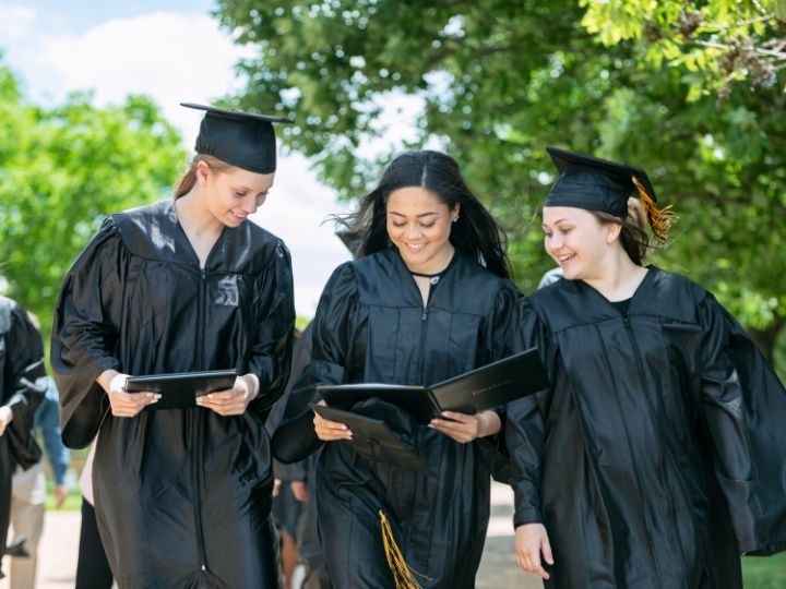 Three graduates in black caps and gowns looking over the details of the middle graduate's diploma after receiving them