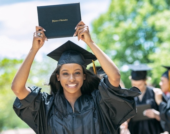 A happy graduate in black cap and gown lifting their diploma up over their head, smiling in excitement