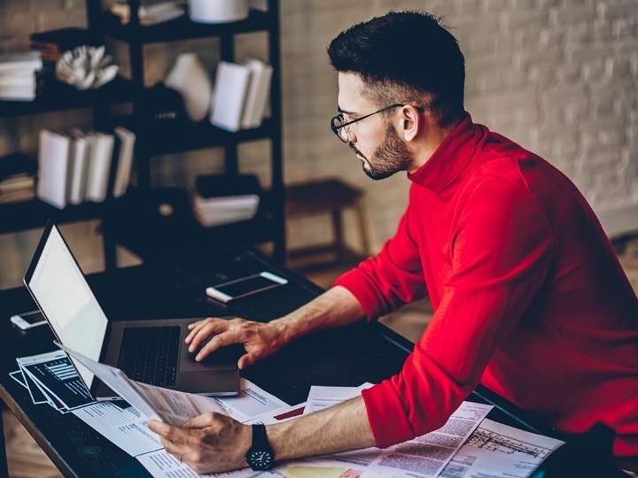 A person going over a project proposal on their laptop at a coffee table covered in paperwork