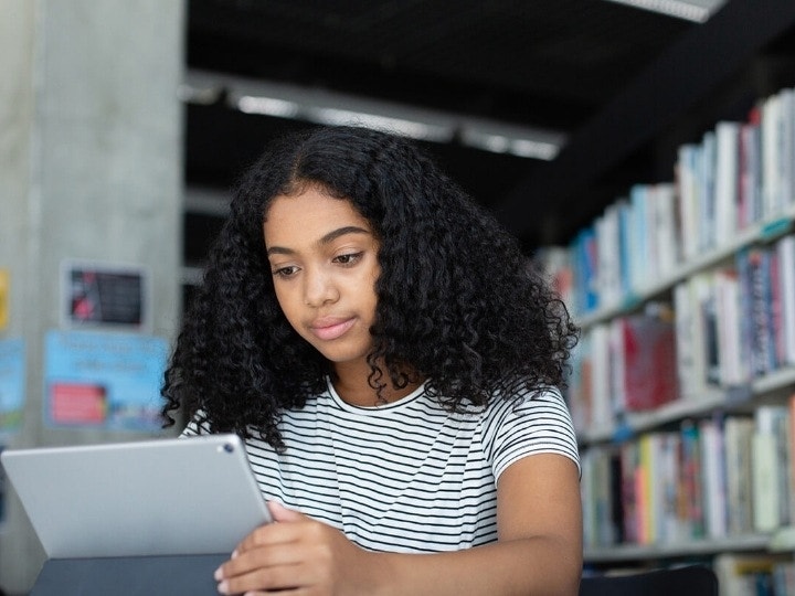 A young student sitting in a library and reading a free eBook on a tablet