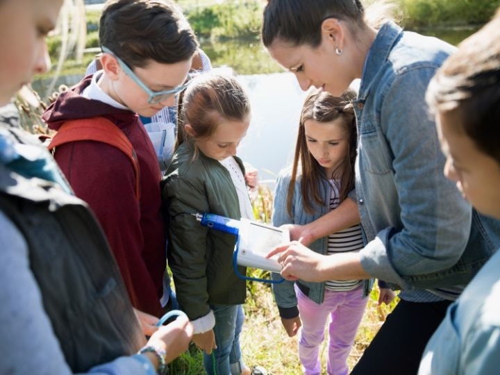 A teacher surrounded by students and showing them something on a tablet-like scientific device during a school field trip