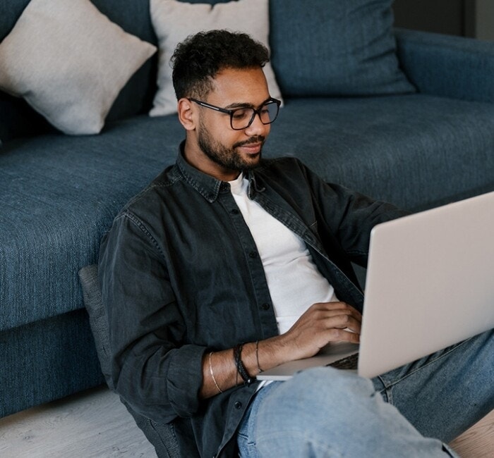 A college student sitting on the floor in front of their couch and using their laptop