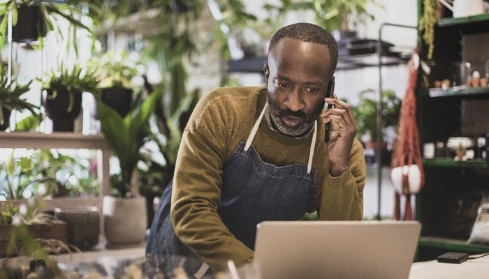 A photo of a plant store owner talking on the phone while looking down at their laptop on a table