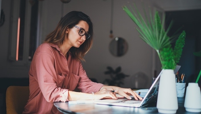 A person sitting at a desk with green foliage, working on a laptop