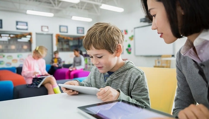 A teacher looking on as a young student reads an eBook on a tablet