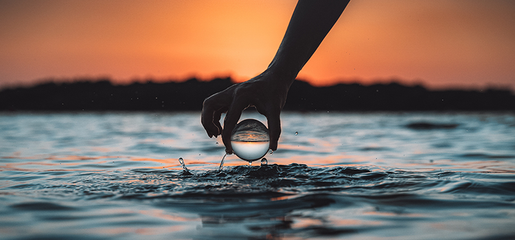 A person holding a lensball above water with a hillside in the far background