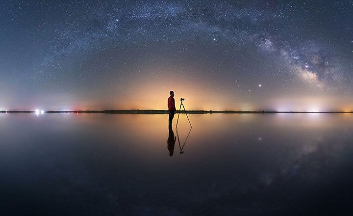 A photographer standing next to a camera on a tripod with the Milky Way seen in the background sky