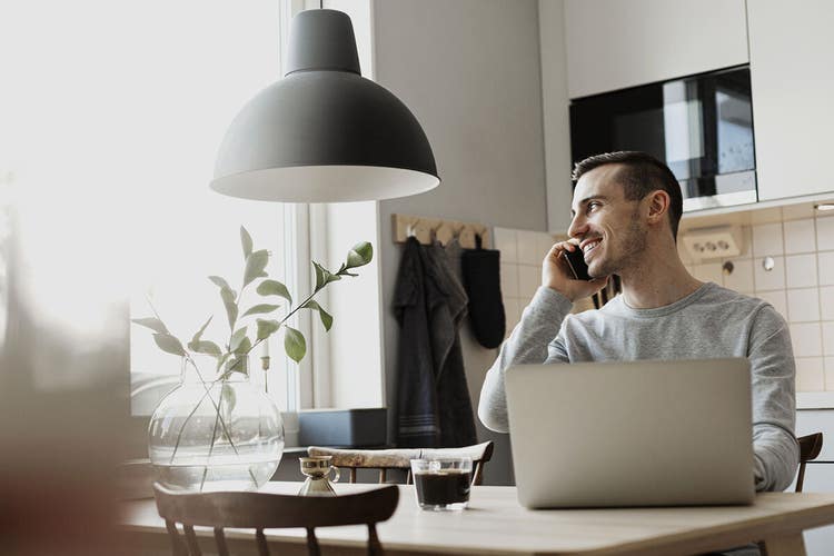 A person talking on the phone while sitting at a table with a laptop and a cup of coffee