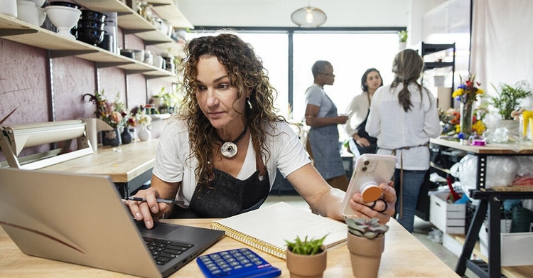 A photo of a florist in a flower shop working at a desk with a laptop, notebook, calculator, and smartphone.