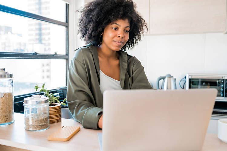 A photo of a person sitting at a counter in a kitchen and working on a laptop computer.