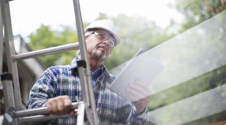 A roof contractor holding a tablet device while standing on a ladder