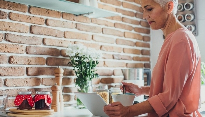 A person standing at a kitchen counter eating from a bowl while reading their tablet