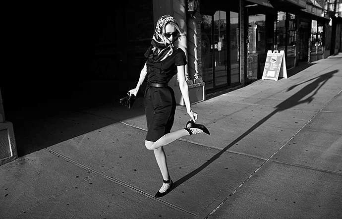 A black-and-white photo capturing a person and their shadow on a sidewalk on top