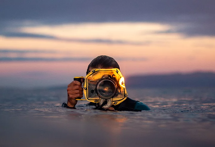 A photographer in a body of water holding their camera equipped with a waterproof housing