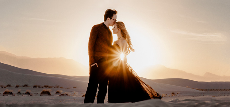 A couple wearing wedding outfits posing in front of a sunset in a desert as a lens flare shines between them